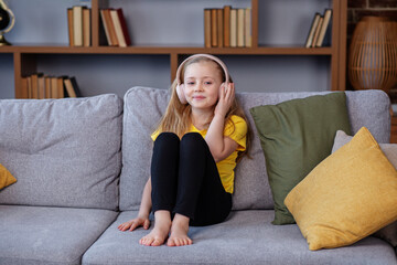 Happy child listens to music or audiobook at home on couch. Little school kid wearing headphones