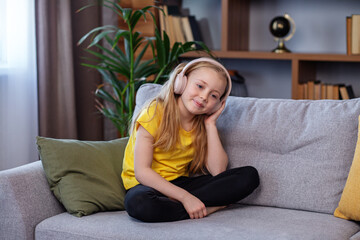 Happy child listens to music or audiobook at home on couch. Little school kid wearing headphones