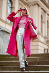 Fashionable confident woman wearing fuchsia color midi coat, sunglasses, metallic pants, combat...