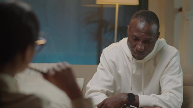 Over the shoulder shot of black man sitting on sofa and having conversation with female psychologist during therapy session in evening