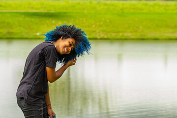 Uma jovem negra, feliz, num parque natural de muito verde, alisando os cabelos tingidos de azul.