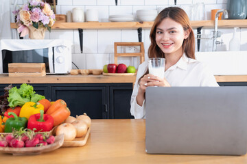 Woman with a beautiful face in a white shirt is making a healthy breakfast with bread, vegetables, fruit and milk inside the kitchen and opening her laptop for cooking lessons. healthy cooking ideas.