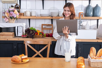 Woman with a beautiful face in a white shirt is making a healthy breakfast with bread, vegetables, fruit and milk inside the kitchen and opening her laptop for cooking lessons. healthy cooking ideas.