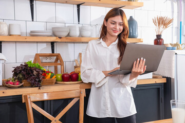 Woman with a beautiful face in a white shirt is making a healthy breakfast with bread, vegetables, fruit and milk inside the kitchen and opening her laptop for cooking lessons. healthy cooking ideas.