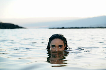 Mystery woman swimming in the ocean , looking at the camera with her big beautiful eyes.  Portrait of calm girl. - 586974828