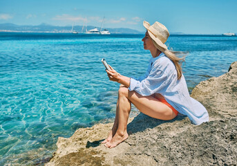 Woman relaxing outdoors lifestyle watching, reading on tablet ebook on the beach in summer day. Wearing wide brimmed hat, Concept of beach vacation.
