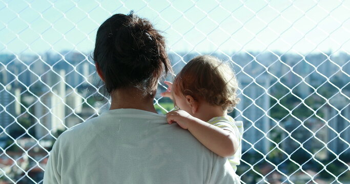Mother holding baby overlooking city at apartment balcony with safety net window