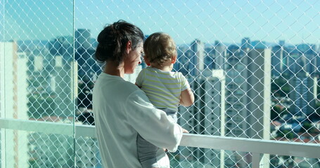 Mom holding baby looking at city view from apartment balcony with safety net - obrazy, fototapety, plakaty