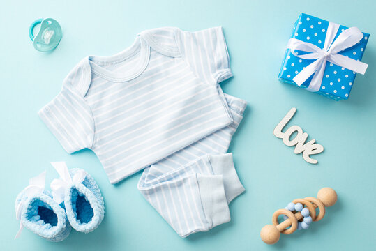 Baby shower concept. Top view photo of infant clothes blue shirt pants inscription love knitted booties giftbox with bow wooden rattle and pacifier on isolated pastel blue background
