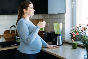 Iron deficiency, anemia, iron boosting smoothie recipes for pregnant woman. Pregnant woman preparing green vitamin smoothie with a blender and drinking in the kitchen.