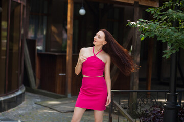 A stylish slender smiling woman with long dark hair, in a pink short sexy skirt and a tank top, stands near a modern building on a city street.