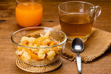 Delicious homemade peach cobbler in transparent glass bowl on bamboo mat with a cup of hot tea on burlap cloth and fresh orange juice in a glass on wooden table.