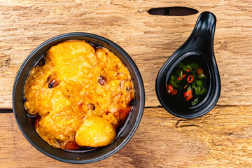 Top view of delicious homemade spicy chicken curry with potato, coconut milk and chilli in black ceramic bowl with fish sauce with red and green pieces of chilli in small black cup on wooden table.