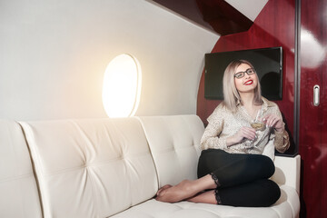 Business pretty lady drinking champagne in luxury private jet, sitting on sofa. Businesswoman traveling in airplane first class. Concept quality of service, travel at highest level. Copy ad text space