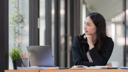 Obraz premium Portrait of an Asian businesswoman thinking and looking away while working with a laptop in a modern office.