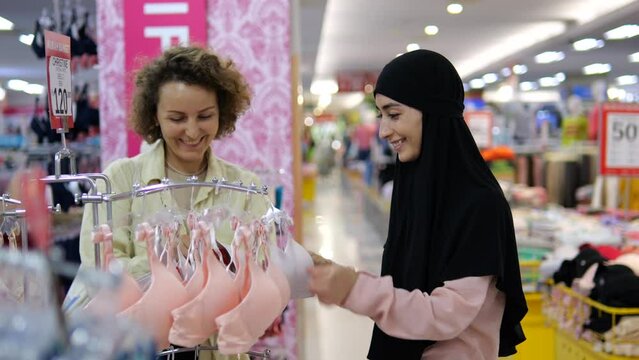 Best friends of different races and religions in clothing store choose underwear and bras, chatting, laughing, smiling. Caucasian and arab woman in hijab on shopping in shopping mall choose underwear