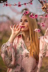 Fototapeta na wymiar The portrait of young sensual blondie woman in pink silk skirt posing holding in teeth the peach blossom into the garden