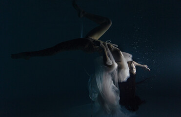 Underwater shoot of beautiful woman in white flying transparent dress dancing in water in sunbeams.