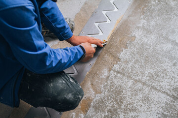 The worker is shown wearing protective gear and kneeling down on the concrete floor, using specialized tools to carefully measure and cut the material to fit precisely.
