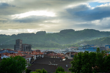 Tiled roofs of an old Spanish town against background of green hills in dawn fog. View of the tower of St. Peter the Apostole Parish in Zumaia