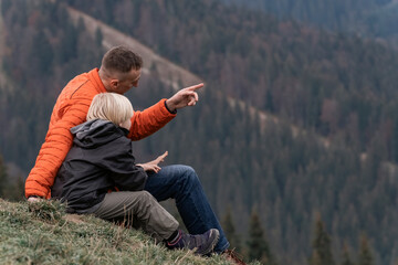 Naklejka na ściany i meble Father and son sit on hillside on the forest background. Trekking and hiking with children. Weekend hike in mountains.