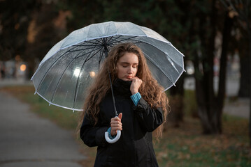 Brooding girl with long hair walks through autumn park in rainy weather under transparent umbrella.