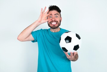 Young man holding a ball over white background keeping eyes opened to find a success opportunity.