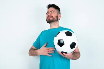 Young man holding a ball over white background touches tummy, smiles gently, eating and satisfaction concept.