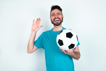 Young man holding a ball over white background Waiving saying hello happy and smiling, friendly welcome gesture.