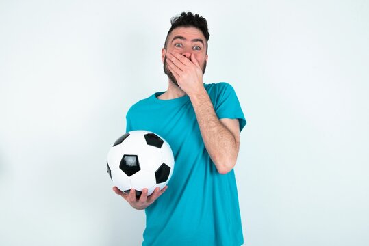 Vivacious Young man holding a ball over white background , giggles joyfully, covers mouth, has natural laughter, hears positive story or funny anecdote
