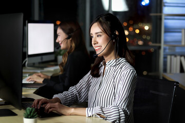 Beautiful Asian woman with headphones work night shift at call center customer care service desk, looking at desktop computer, typing answer question, female with hands-free phone work hard overtime.