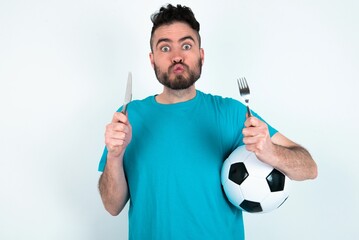 hungry Young man holding a ball over white background holding in hand fork knife want tasty yummy...