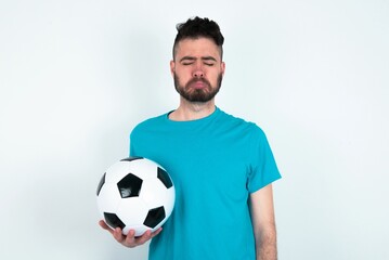 Dismal gloomy rejected Young man holding a ball over white background has problems and difficulties, curves lower lip and closes eyes in despair, being in depression