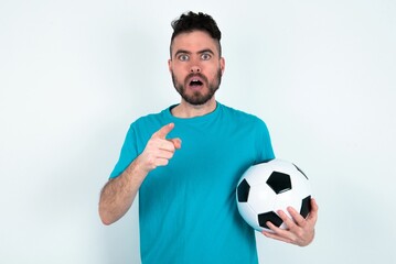 Naklejka premium Shocked Young man holding a ball over white background points at you with stunned expression