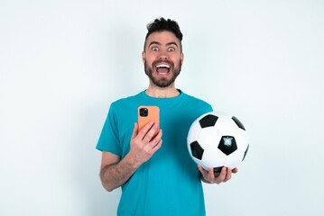Young man holding a ball over white background holds mobile phone in hands and rejoices positive news, uses modern cellular