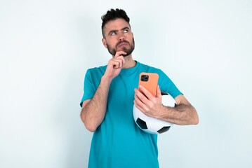 Young man holding a ball over white background thinks deeply about something, uses modern mobile...