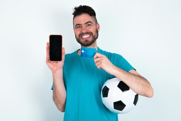 Photo of adorable Young man holding a ball over white background holding credit card and smartphone. Reserved for online purchases
