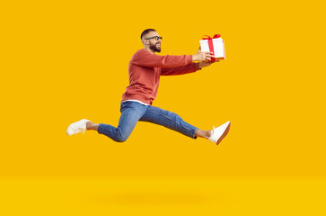 Fototapeta na wymiar Charming smiling young man in glasses wearing red sweatshirt and jeans jumping high holding gift smiling isolated on yellow background. Holiday gifts, discounts, sales, marketing, good offer concept.