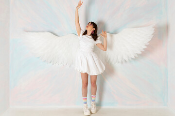 Cute like an angel brunette girl in a fancy short white dress, huge feather wings and bright makeup agains a sky background.