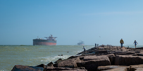 The South Jetty concrete sidewalk, with people fishing and walking, and oil tanker ships approaching from the Gulf of Mexico on the channel to Corpus Christi, Texas. 