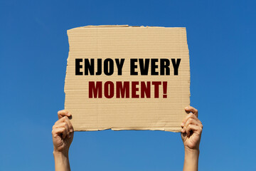 Enjoy every moment text on box paper held by 2 hands with isolated blue sky background. This...