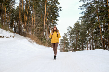 Young woman traveler with  backpack and yellow coat walking on snow covered road in winter forest in frosty weather.