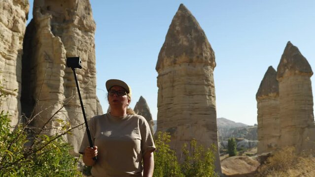 Caucasian girl blogger in glasses and a yellow baseball cap is recording a story on an action camera. Against the background of a sandstone rock in the form of phalos. Valley of Love in Cappadocia