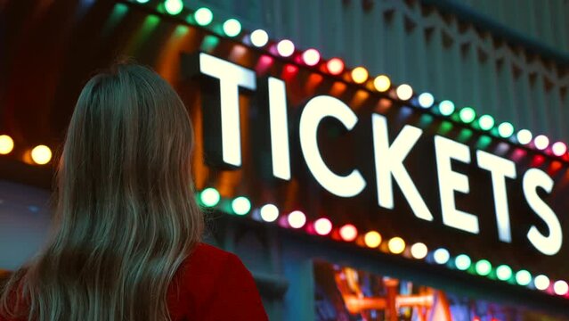 Young woman smiles at vintage carnival ticket sign at amusement park. Back view of female person enjoying evening fun, bright lights of fairground in background.