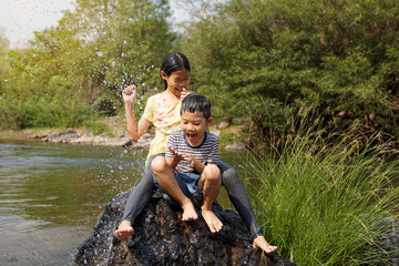 Asian girls and boys take a break from playing in the river to sit on the rocks. Both of them laughed and dodged the water that had been poured over them. Water droplets spread and look fresh.   - obrazy, fototapety, plakaty