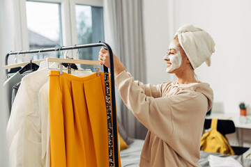 A happy Caucasian woman with a mask on her face is choosing clothes to wear