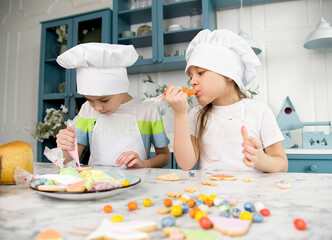 two little cute funny smiling kids wearing a white chef's hats and uniform decorating easter bread with food coloring  in the blue and white kitchen with easter decorations