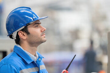 Portrait of a white European male engineer side view looking at the destination with determined eyes holding a radio in an industrial factory Wear a helmet and uniform.