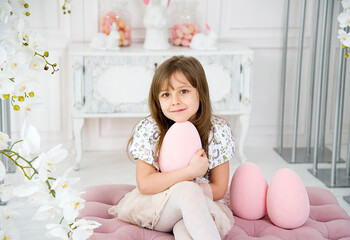 little funny cute smiling brunette girl sitting on зink sofa and playing with three big pink toy easter egg  in room with white orchids