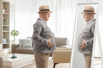 Naklejka na ściany i meble Elderly man with a big belly looking at a mirror at home